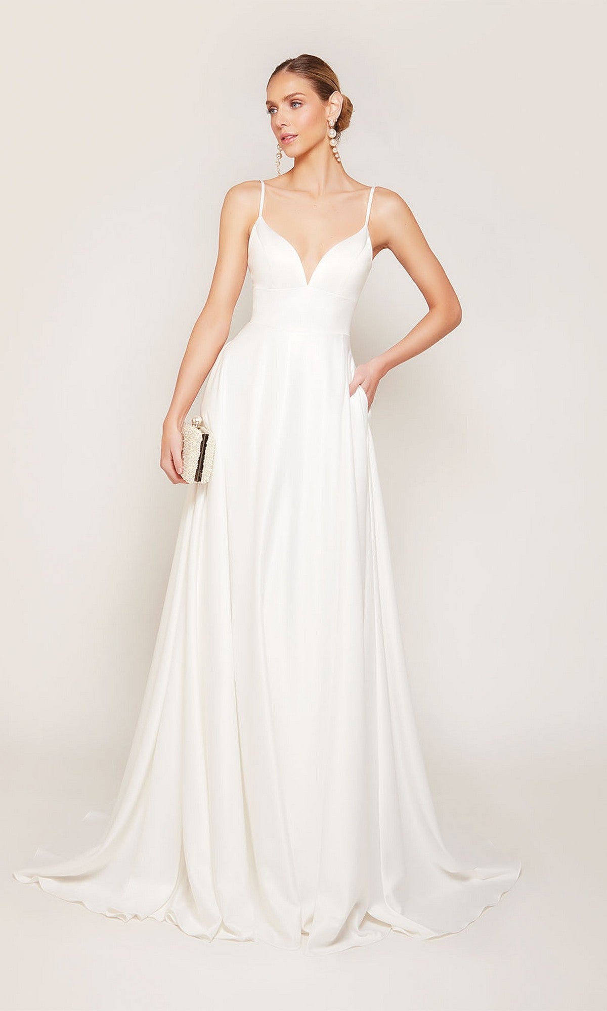 A woman stands against a plain light background wearing the Alyce 7095 long white dress, featuring satin fabric, thin straps, and a V-neckline. She holds a small clutch in one hand.